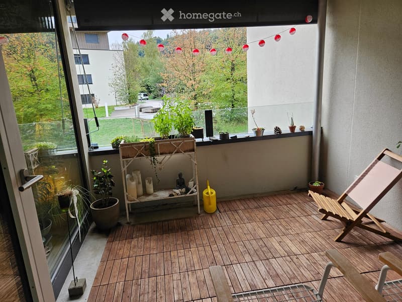 Balcony with wooden decking, potted plants, a small shelf with candles and a statue, a yellow watering can, and a wooden folding chair.