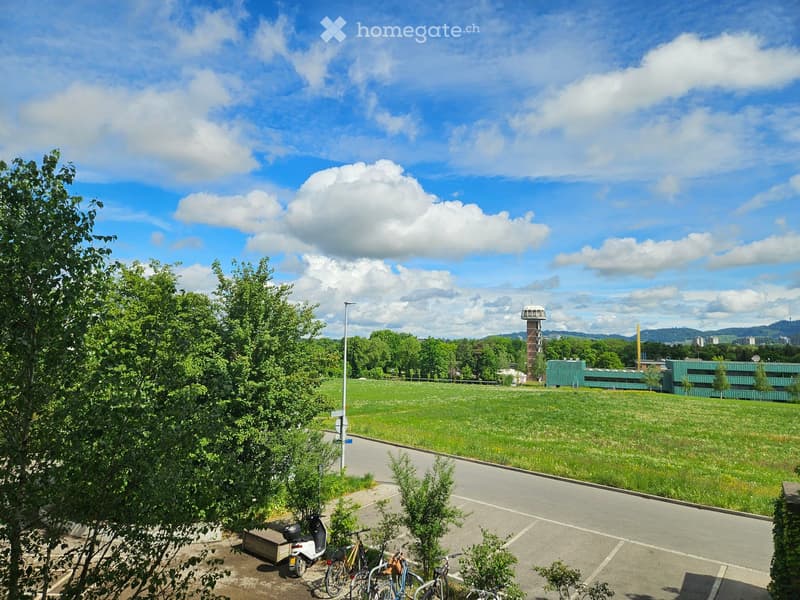 A grassy field with trees and a modern building in the background, under a partly cloudy sky. There are parked bicycles and a scooter in the foreground.