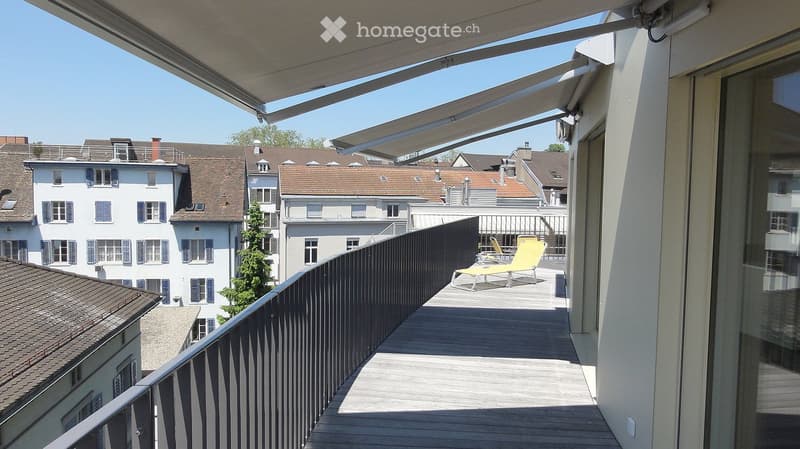 Balcony with wooden decking, black railing, sun lounger, and retractable awning, overlooking a cityscape with traditional houses.