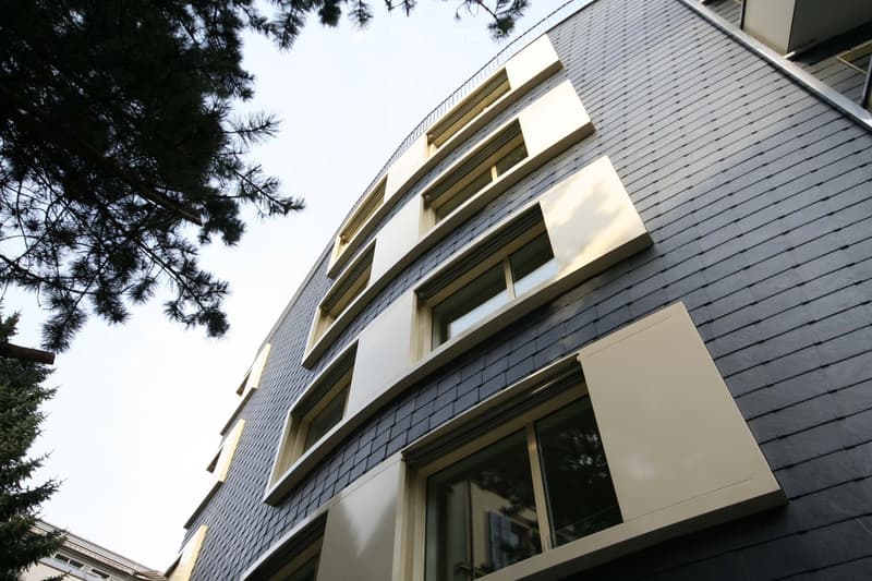 Modern multi-story building with dark grey shingled exterior, large windows with white frames, and light-colored shutters.