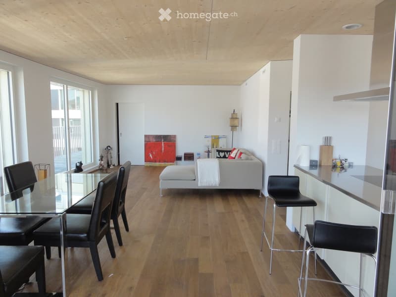 Modern living room with a white sofa, a glass dining table, black chairs, and bar stools. The room features wooden flooring, large windows, and a kitchen area with white cabinets.
