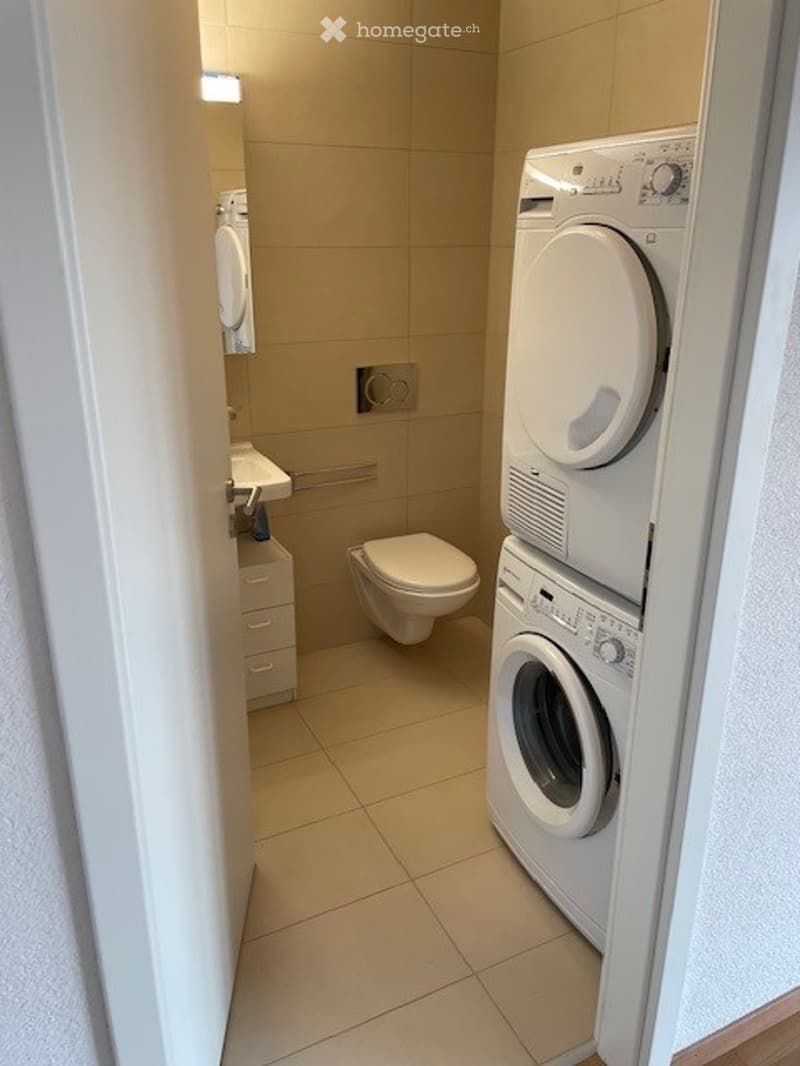 Modern bathroom with beige tiles, a toilet, sink, mirror, cabinet, and stacked washing machine and dryer.
