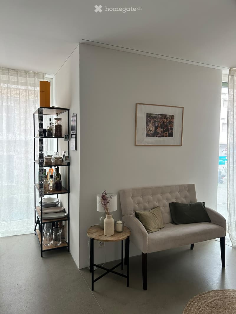 Modern living room with a grey tufted sofa, wooden side table, decorative vase, shelf with various items, and a framed picture on the wall.