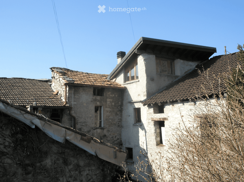 Two-story stone house with a tiled roof, multiple windows, and visible wear and tear.