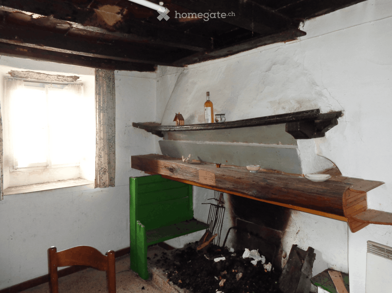 An empty room with exposed wooden beams, a window, shelves, and a fireplace with debris. No furniture or equipment is present.