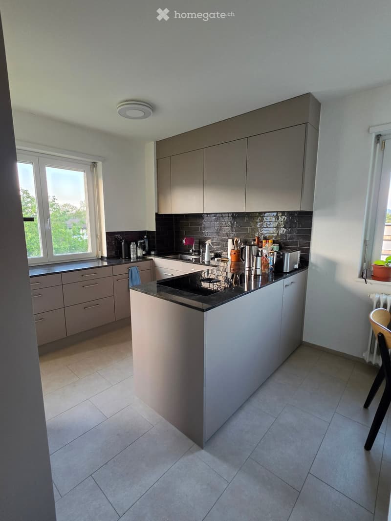 Modern kitchen with grey cabinets, black countertop, and stainless steel appliances. Features a central island with sink and faucet, and various kitchen items on the counter.