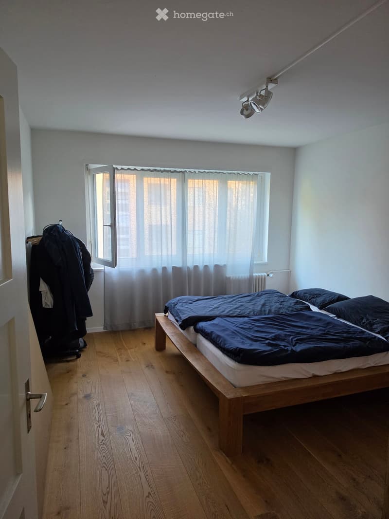 A modern bedroom with wooden flooring, a single bed with dark blue bedding, a window with white curtains, a radiator, and a wardrobe with hanging clothes.