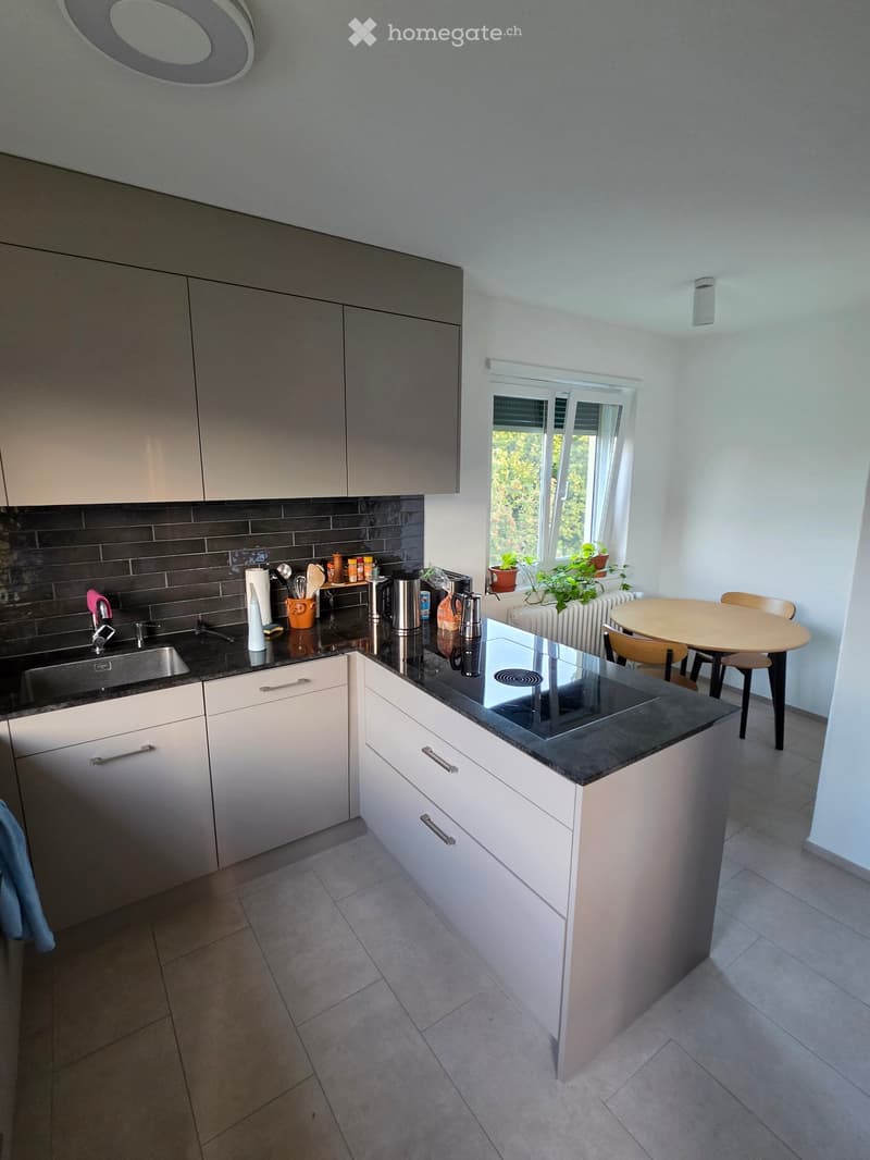 Modern kitchen with white cabinets, black countertop, induction stove, sink, and small dining area with table and chairs. Plants on windowsill.