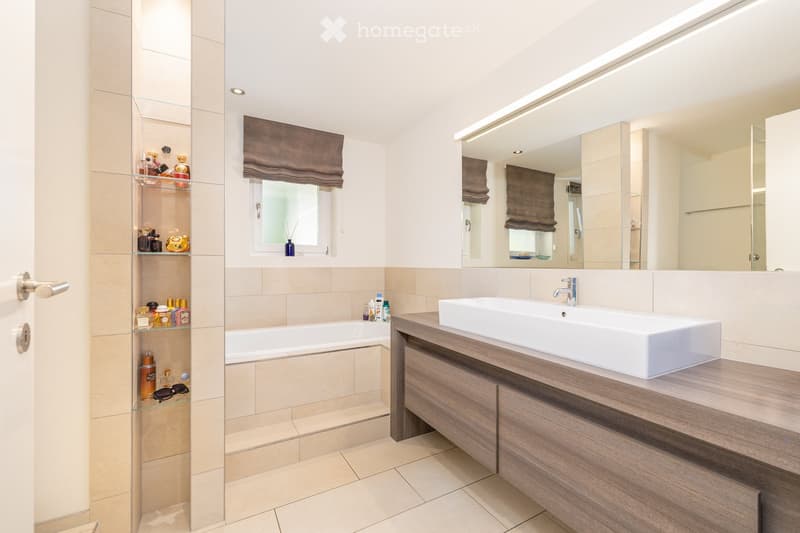 Modern bathroom with a large bathtub, a white sink on a wooden vanity, a large mirror, and a glass shelf holding toiletries.