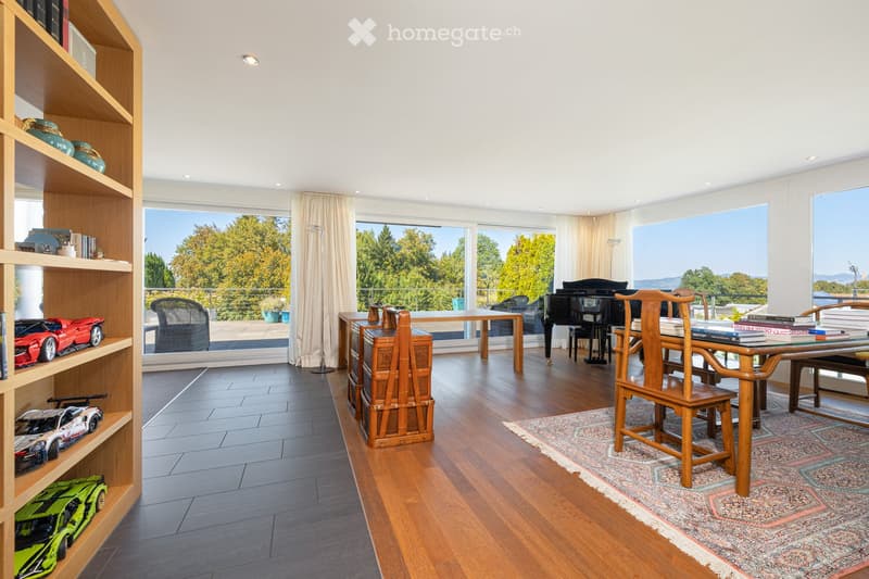 A spacious, well-lit room with wooden flooring, large windows offering a scenic view, a wooden shelf with decorative items and toy cars, a piano, a dining table with chairs, and a patterned rug.
