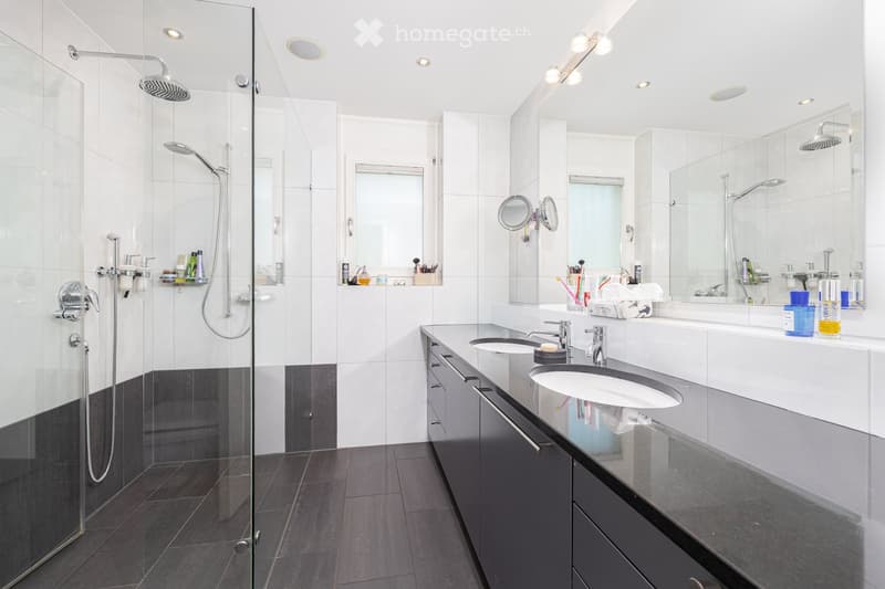 Modern bathroom with two sinks, a large mirror, a glass-enclosed shower, and dark grey tiles on the floor and lower walls.