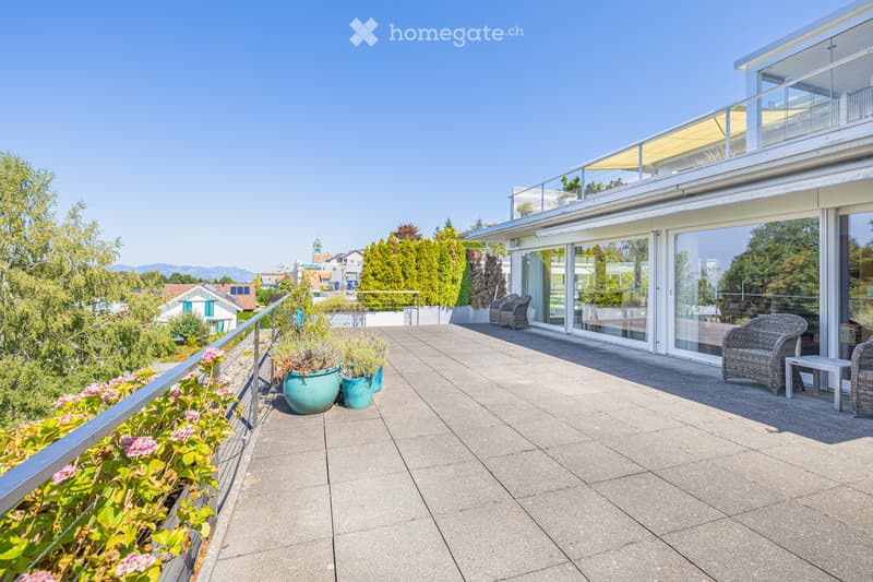 Balcony with modern glass railings, outdoor seating, and potted plants, overlooking a scenic landscape with trees and distant buildings.