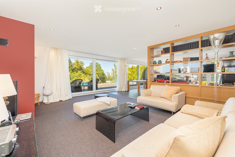 Modern living room with large windows, cream-colored sofa, coffee table, ottoman, floor lamp, and bookshelf with decorative items.