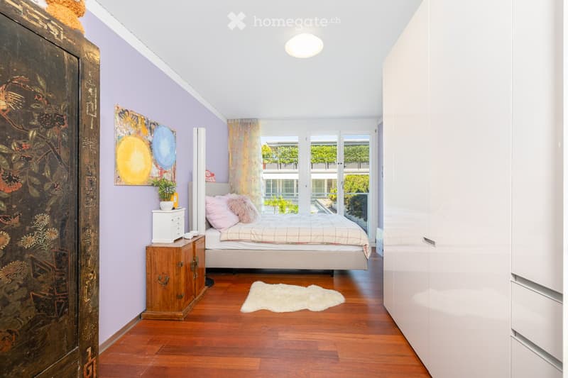 Cozy bedroom with wooden flooring, a bed with pink pillows and a plaid blanket, a nightstand with a plant, a painting on the wall, and a white rug on the floor.