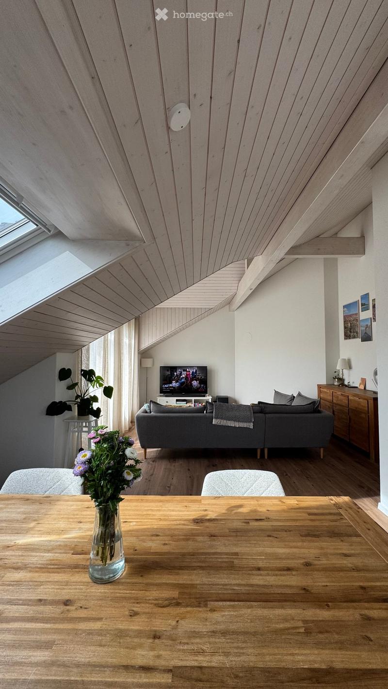 Modern living room with wooden floors, slanted wooden ceiling, large windows, grey sofa, wooden dining table, vase with flowers, and television.