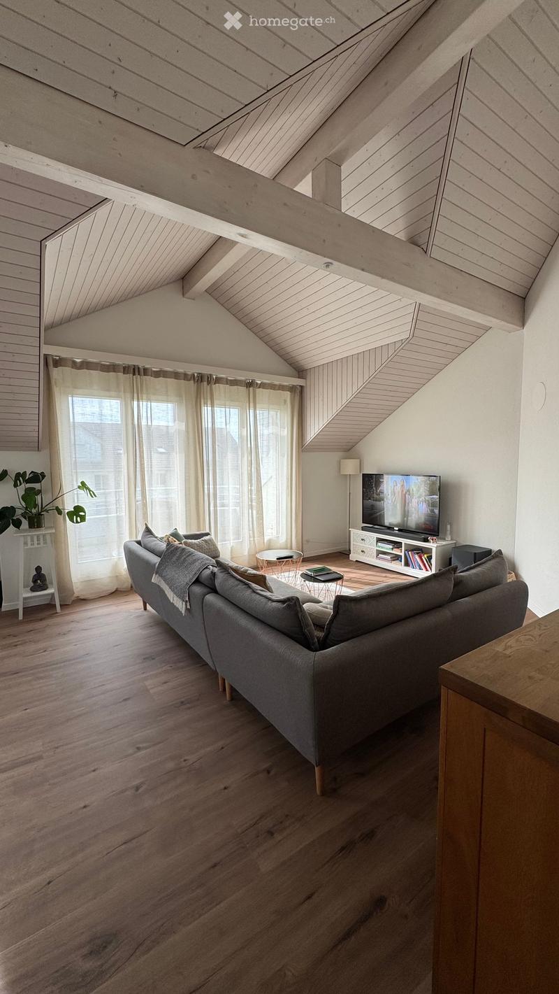 Modern living room with wooden floors, slanted wooden ceiling, large windows with sheer curtains, grey sectional sofa, coffee table, TV on a white stand, small round tables, and a potted plant.