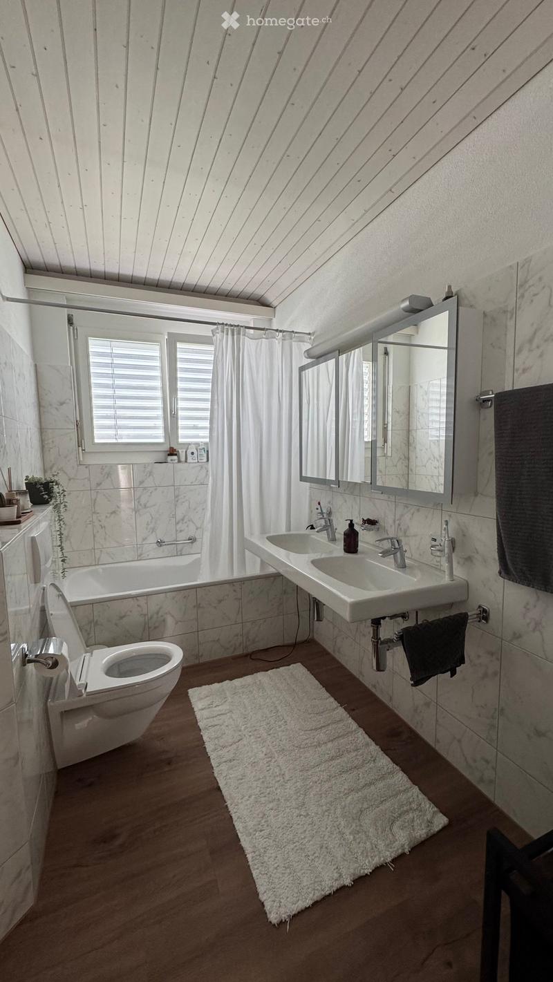 Modern bathroom with marble tiles, double sink, toilet, bathtub, large mirror, and a white shag rug on the floor.