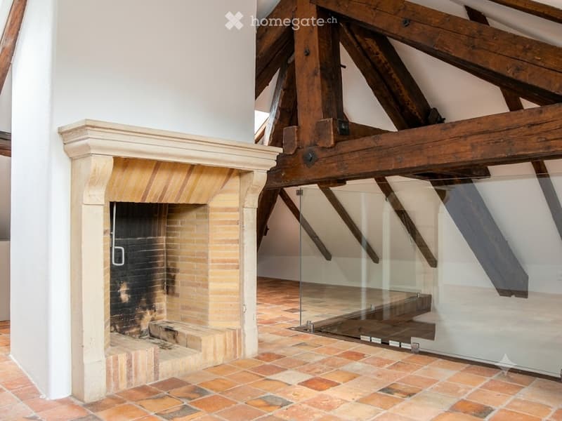 A room with exposed wooden beams and a traditional brick fireplace, featuring a glass staircase leading upwards.