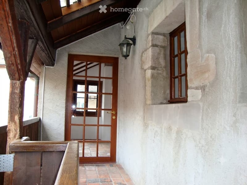 A narrow hallway with exposed wooden beams, a wooden door with glass panels, and a small window on the right side.