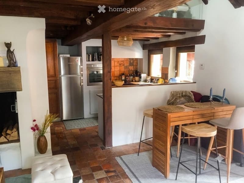 Cozy, rustic kitchen and living area with exposed wooden beams, a fireplace, a refrigerator, an oven, a bar table with stools, and a small seating area.