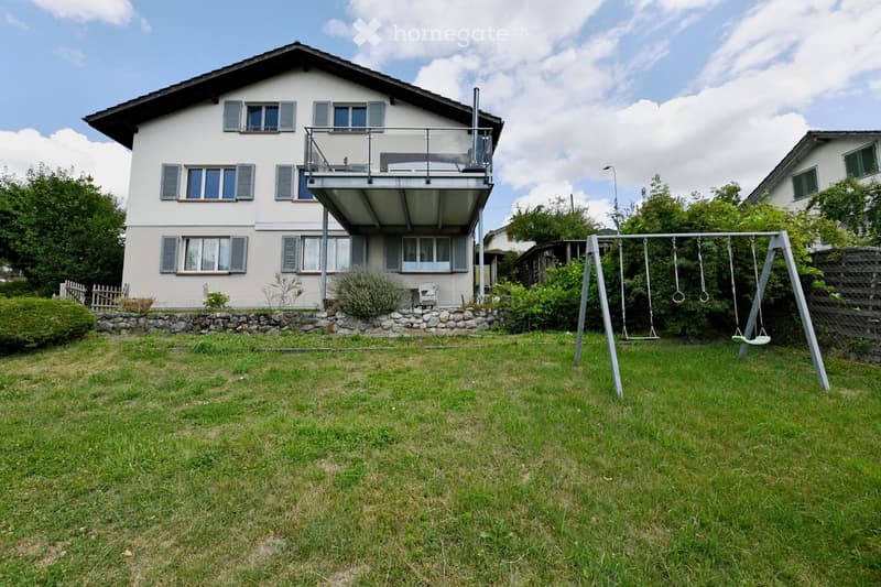 Two-story house with white exterior, multiple windows, and a balcony on the upper floor. The front yard includes a swing set and a small stone retaining wall.