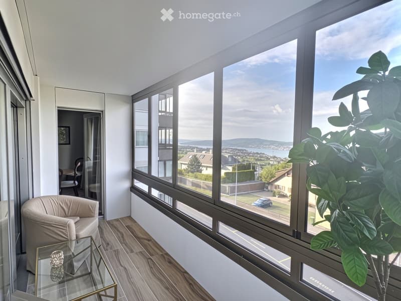 Bright, modern living space with large windows offering a scenic view of the lake and surrounding landscape. Features a light-colored sofa, a glass coffee table, and a potted plant near the window.