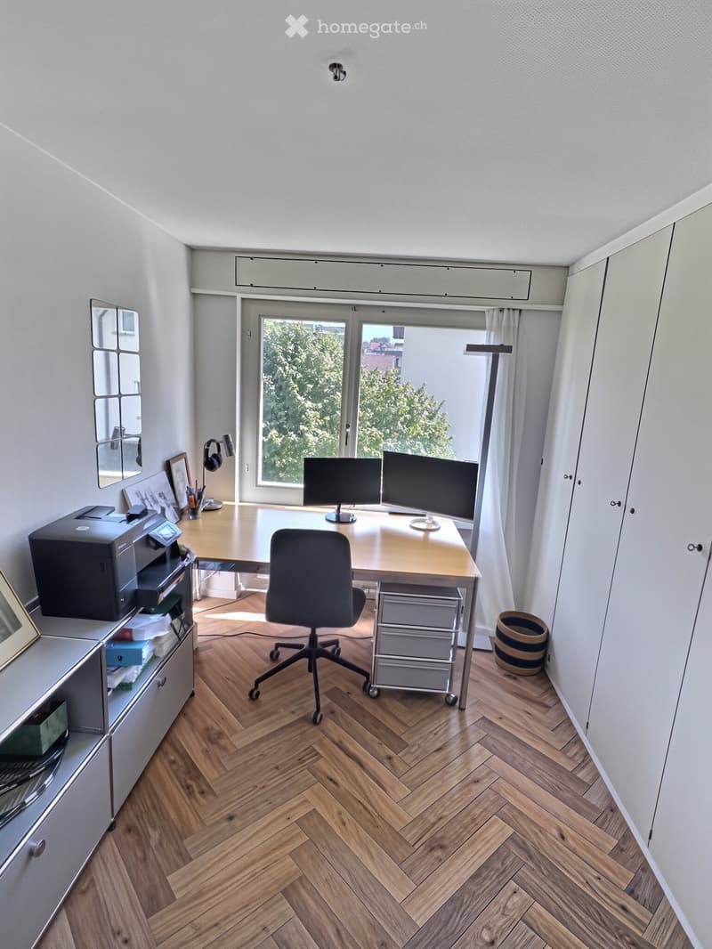 A modern, well-lit office space with a wooden herringbone floor, large window, and white walls. The room features a desk with two monitors, a chair, a printer, and a cabinet with framed pictures and office supplies.