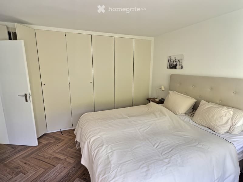 Modern bedroom with a large bed, white bedding, a tall wardrobe, a nightstand with a lamp, and a picture frame on the wall.