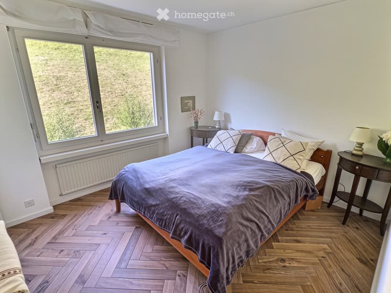 Cozy bedroom with wooden flooring, large windows offering a scenic view, a neatly made bed with pillows and a duvet, two nightstands with lamps, a small table with a vase of flowers, and a picture frame on the wall.