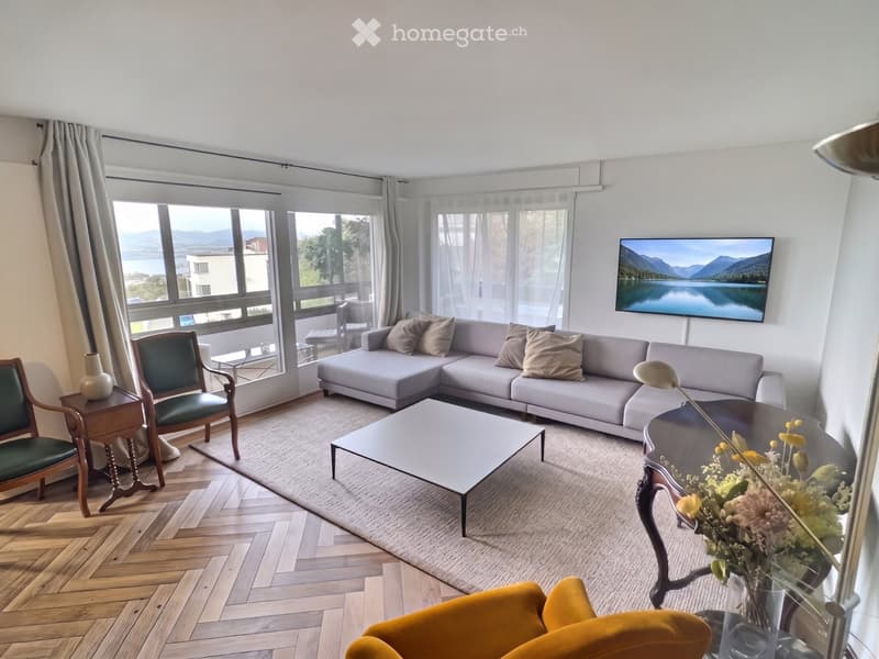 Modern living room with large windows offering a scenic view, featuring a grey sectional sofa, wooden coffee table, elegant armchair, glass side table, decorative vase, and a television displaying a nature scene.