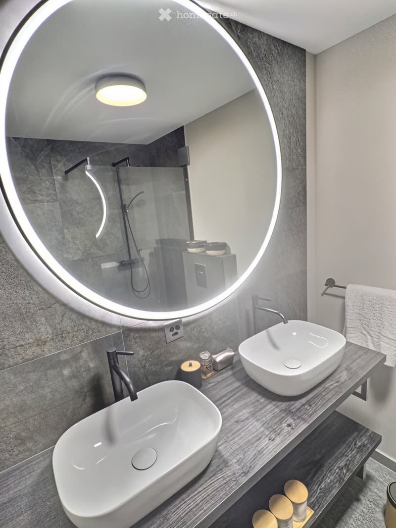 Modern bathroom with two white sinks, sleek faucets, a large round mirror, and a towel on a rack.