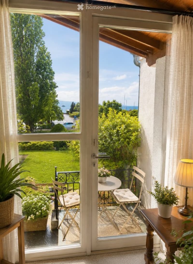 Balcony with a view of a green lawn and trees, featuring a small table, chairs, and potted plants.