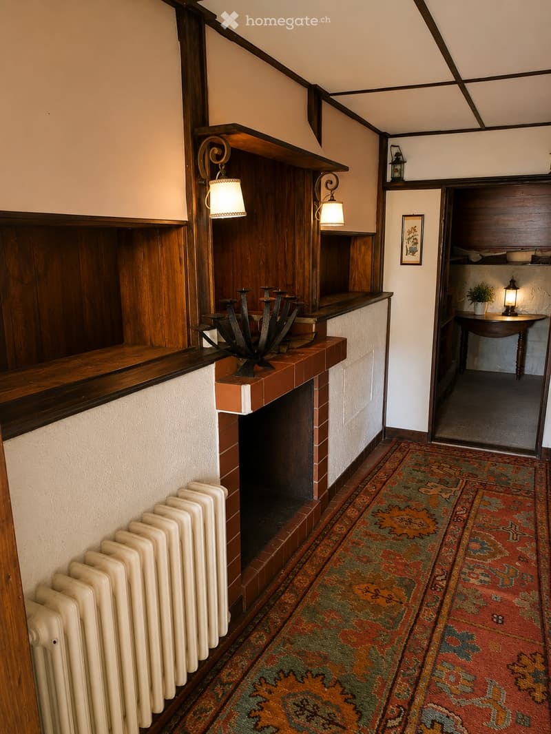 A narrow hallway with wooden paneling, a decorative fireplace, a patterned rug, and a small table with a lamp in the adjacent room.