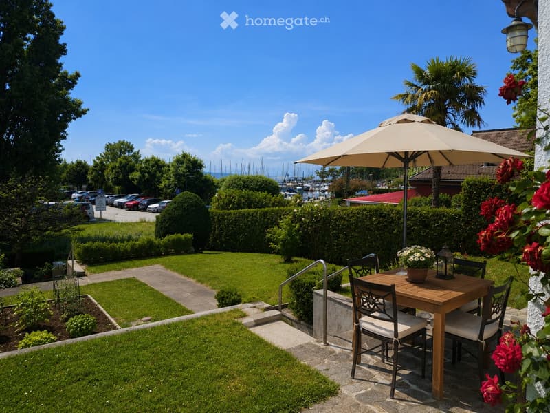 Garden with a wooden table, chairs, umbrella, and flower pot, surrounded by neatly trimmed hedges and a view of a harbor with boats.