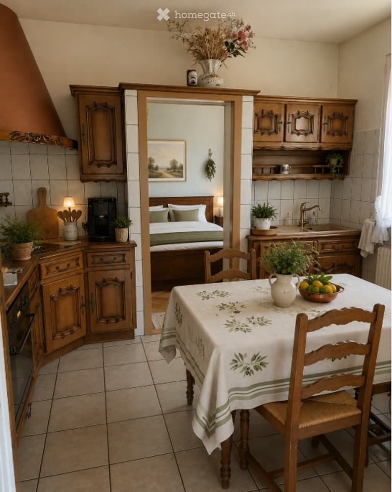 The image shows a cozy kitchen and dining area connected to a bedroom. The kitchen features wooden cabinets, a sink, and an oven. A dining table with a floral tablecloth and chairs is placed in the center. The bedroom visible through an open doorway has a