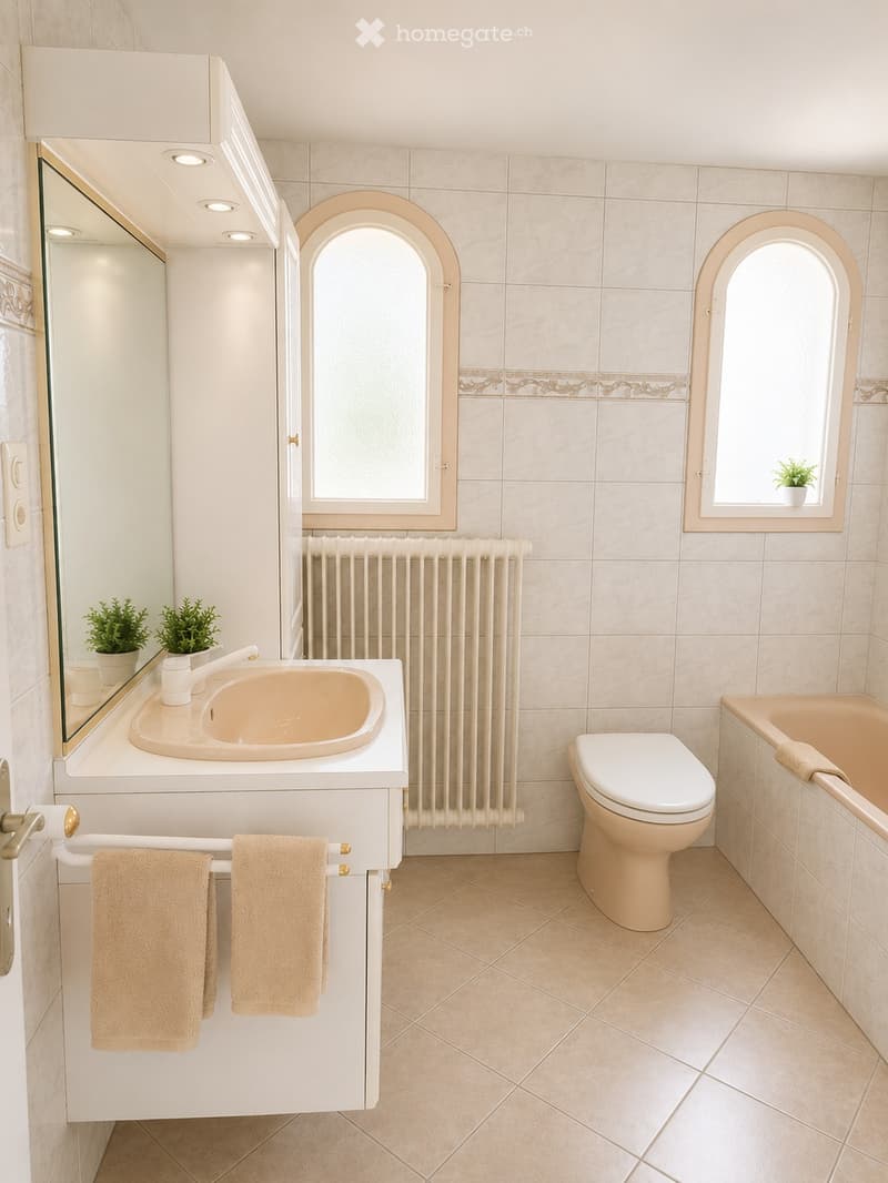 Bathroom with beige tiles, a white sink, a toilet, a bathtub, a radiator, a large mirror, and two small potted plants on the windowsill.
