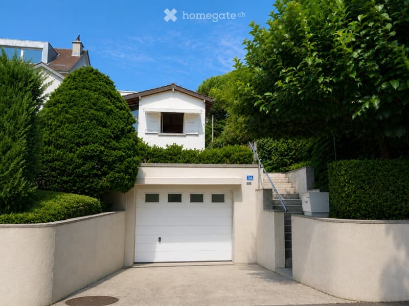 Modern two-story house with a raised ground floor, white garage door, and a staircase leading up to the main entrance.