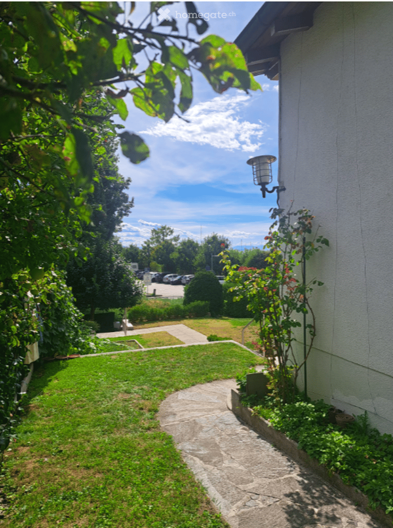 A well-maintained garden area with a stone pathway leading to a house, surrounded by green grass, shrubs, and trees. The background shows a parking area with several cars and a clear blue sky.