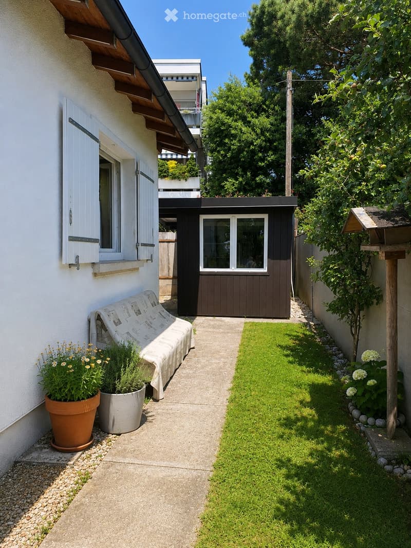 A narrow garden path leads to a small seating area with a covered bench and potted plants, surrounded by greenery and a wooden shed.