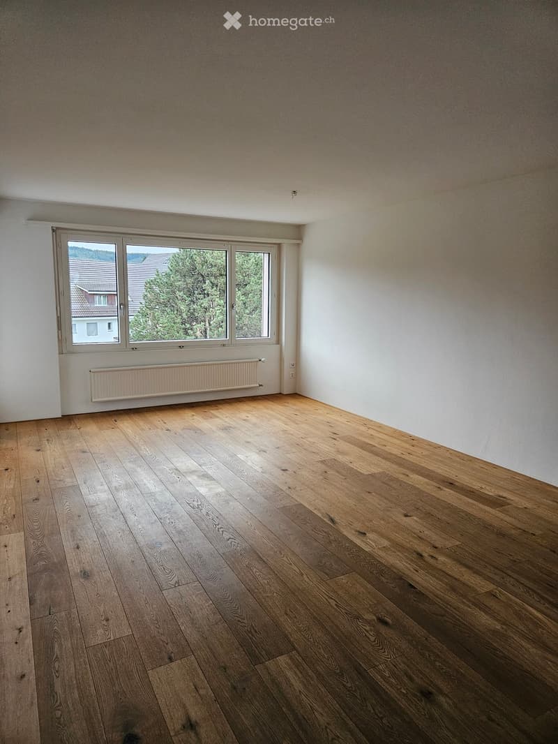Empty room with wooden flooring, white walls, and a large window with a view of trees and a neighboring house.