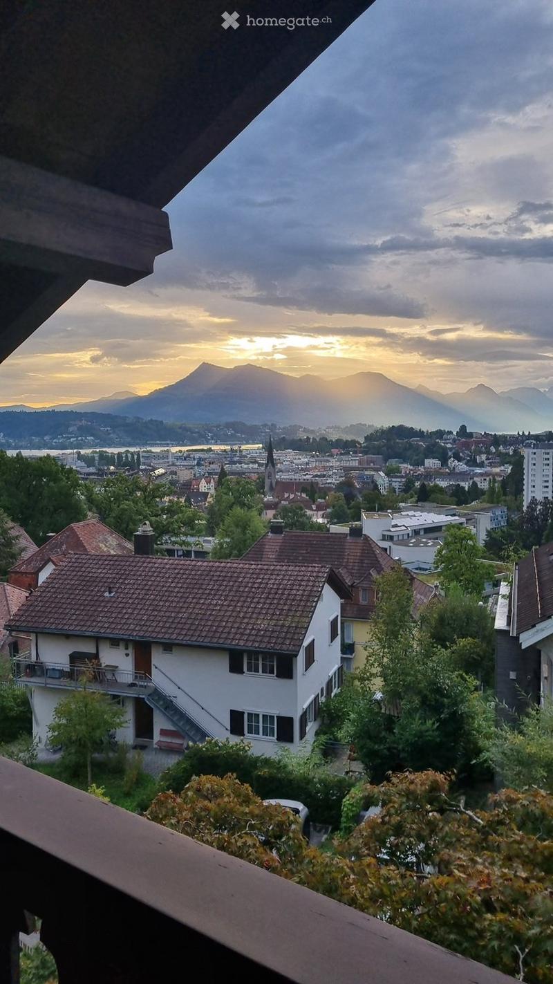 Balcony overlooking a cityscape with mountains in the background, featuring a view of rooftops, trees, and a distant lake.