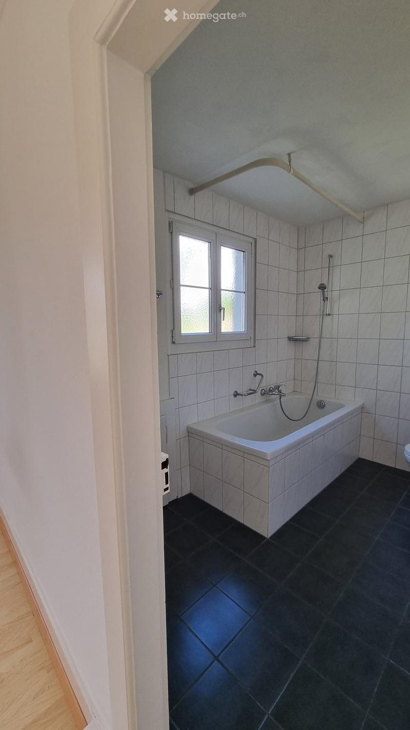 Bathroom with a white bathtub, black tiled floor, grey tiled walls, and a window with frosted glass panes.