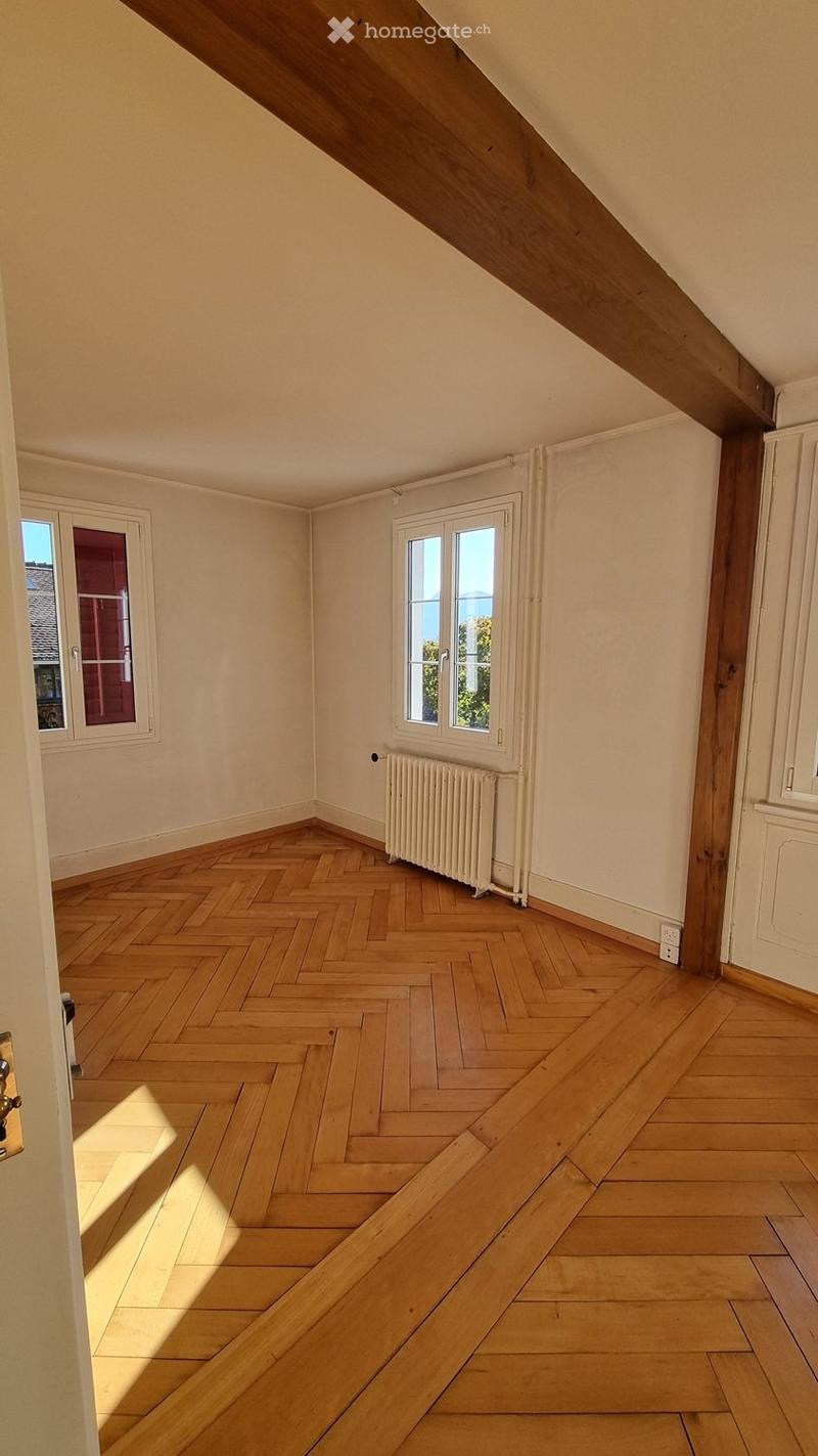 Empty attic room with wooden herringbone flooring, white walls, and large windows allowing natural light. Exposed wooden beams on the ceiling and a radiator under one window.