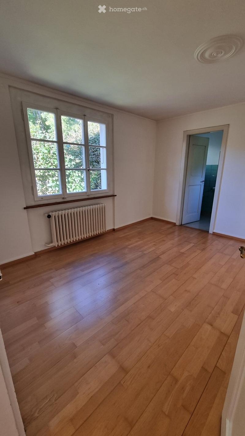 Empty room with wooden flooring, white walls, large windows with white frames, and a radiator below the windows. A door leads to another room.