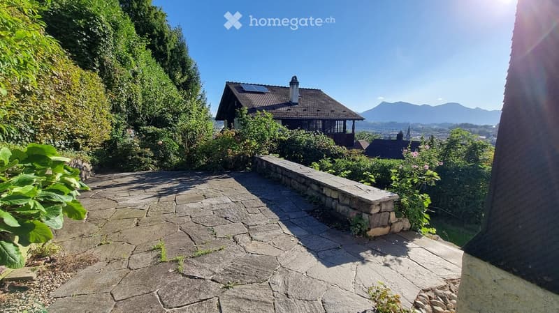 Stone-paved outdoor area with a low stone wall, lush greenery, and a view of a house and distant mountains.