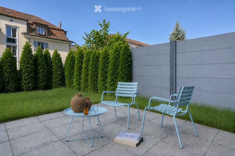 A well-maintained garden area with a light blue metal table and two matching chairs, a small round table with a vase and a cup, and a stack of books on the ground. The garden is enclosed by a grey wooden fence and features neatly trimmed green hedges and