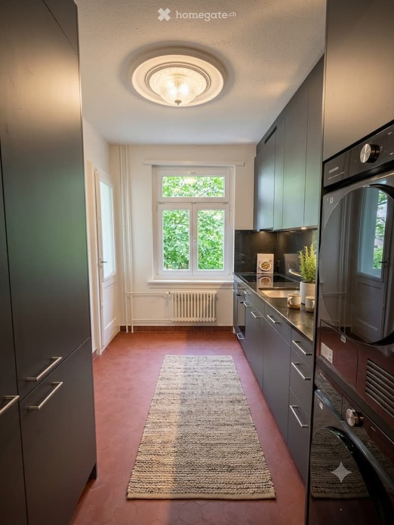 Modern kitchen with dark grey cabinets, stainless steel appliances, a central island, and a large window overlooking greenery. Features a round ceiling light fixture, a radiator below the window, and a textured rug on the terracotta floor.