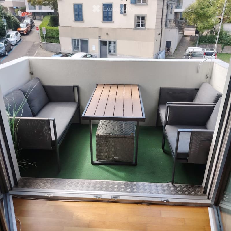 Balcony with green artificial turf, grey sofa, wooden table, and wicker chair.