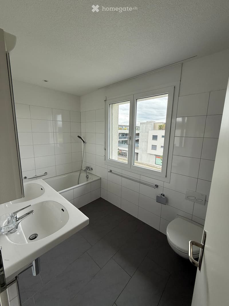 Modern bathroom with white tiles, double sink, bathtub, toilet, and large window offering a view of the city.