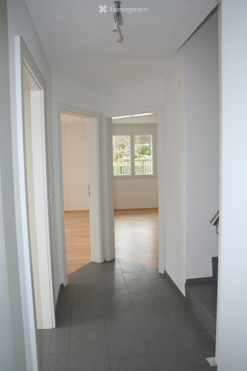 Empty hallway with tiled floor, white walls, wooden flooring in one room, visible windows and railings